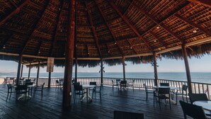 Un área de estar al aire libre con techado, mesas y sillas, con vista a la playa.