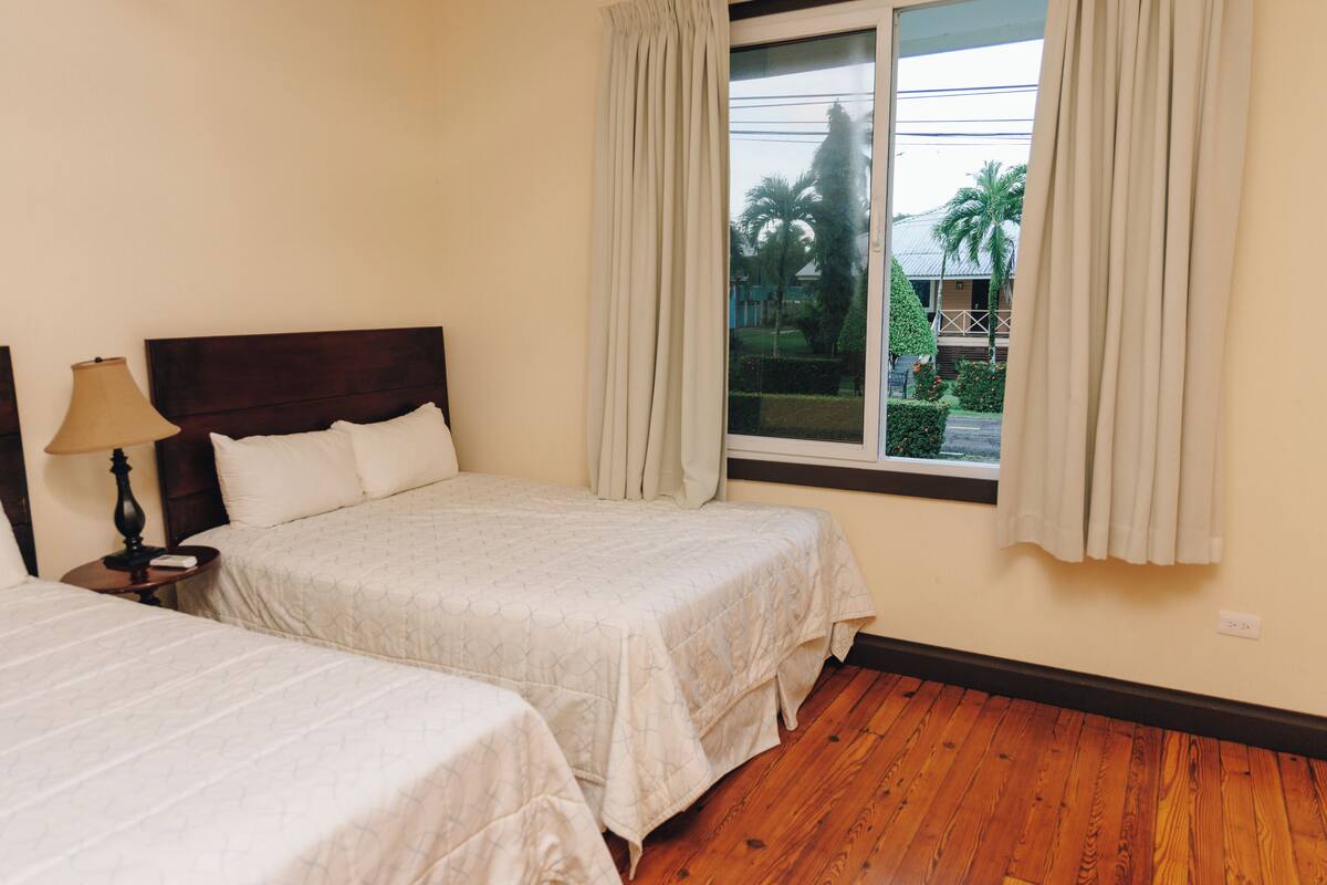 Quarto de hotel com duas camas, uma janela com cortinas, piso de madeira e mesa de cabeceira com abajur.
