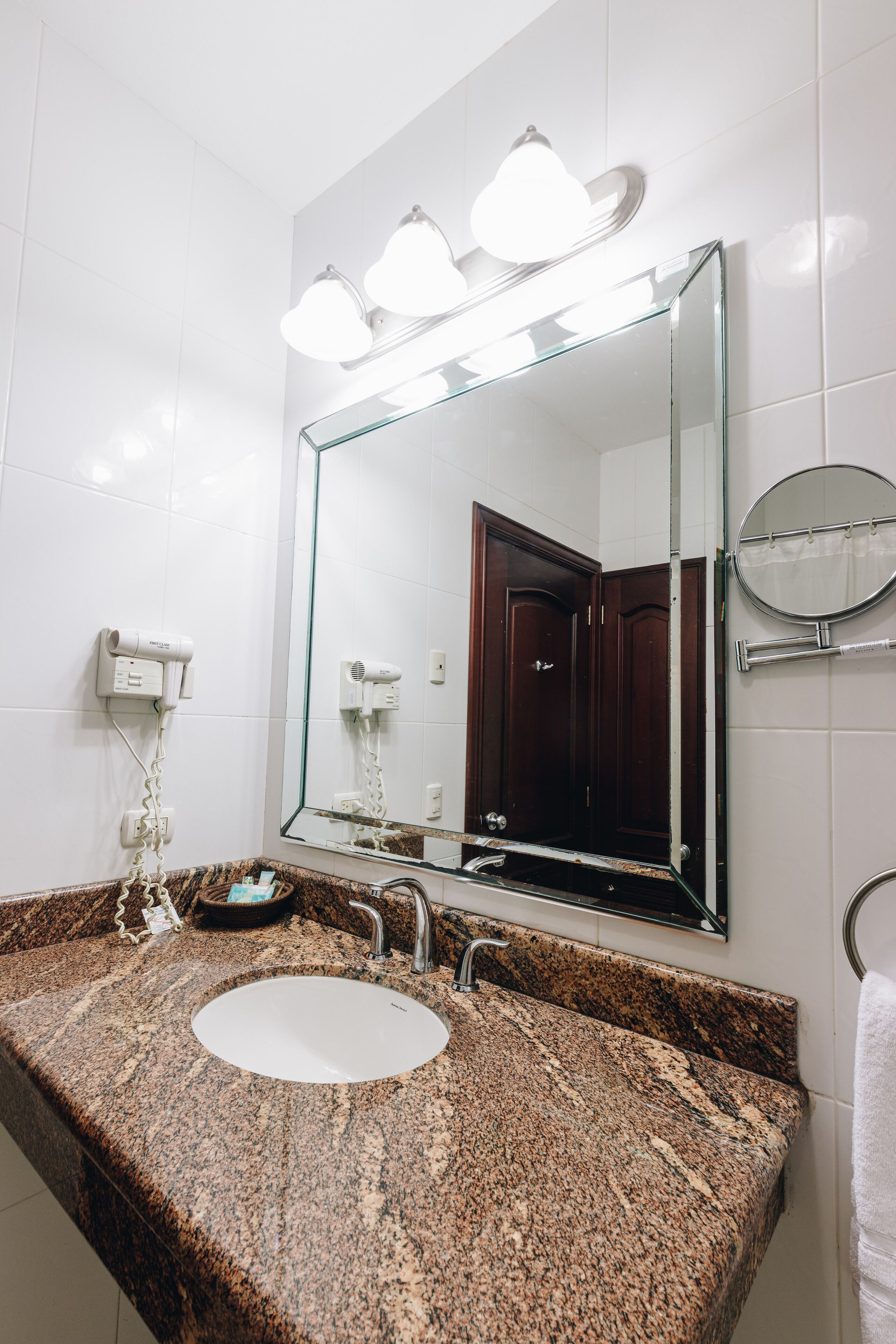 A bathroom with a granite countertop sink, a large mirror with lights, a hairdryer, and a towel rack.