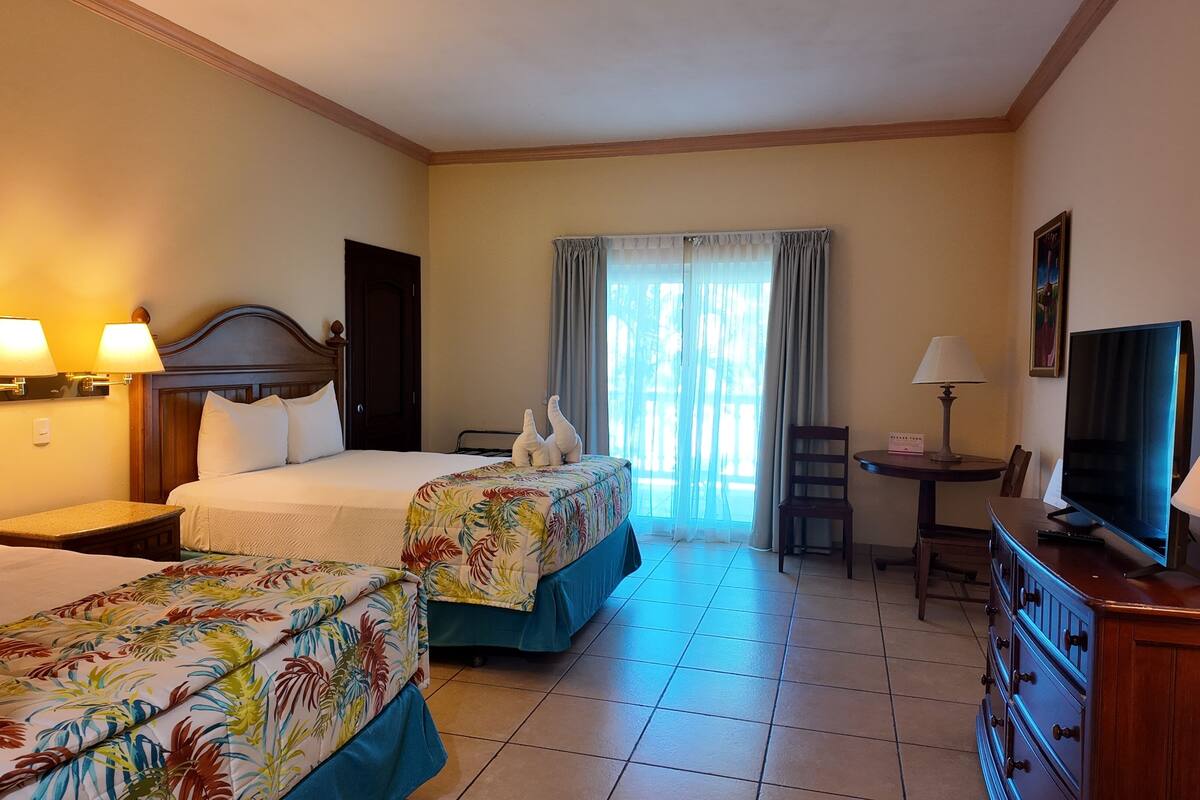 Quarto de hotel com duas camas, uma televisão, uma escrivaninha com abajur e vista para o exterior pela janela.