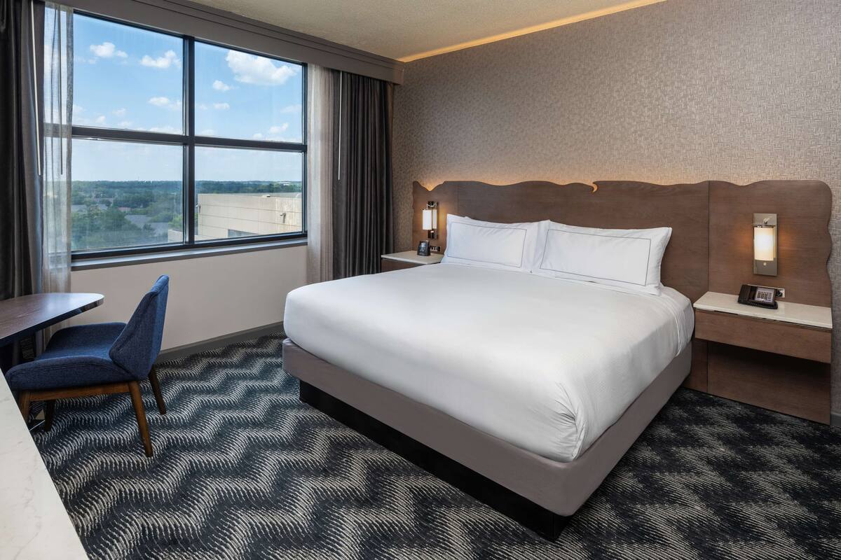 A hotel room with a large bed, a desk, a chair, and a view of the city through the window.