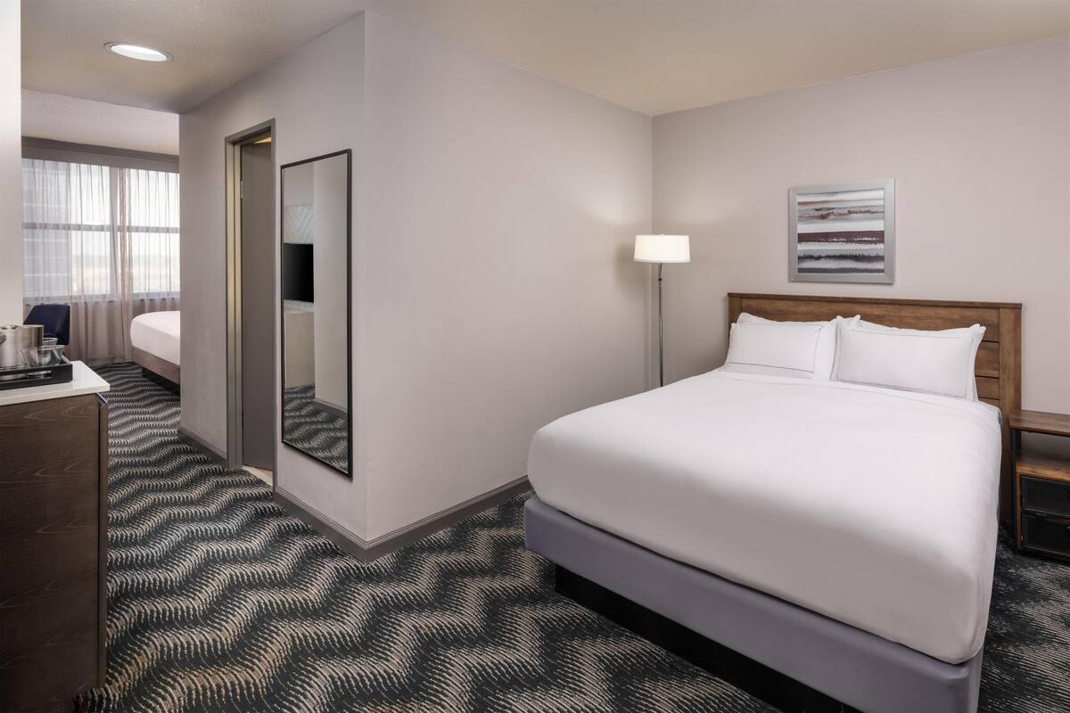 A hotel room with a large bed, a bedside table, a desk with a chair, a mirror, a lamp, and a window with curtains.