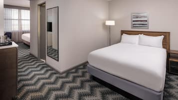 A hotel room with a large bed, a bedside table, a desk with a chair, a mirror, a lamp, and a window with curtains.