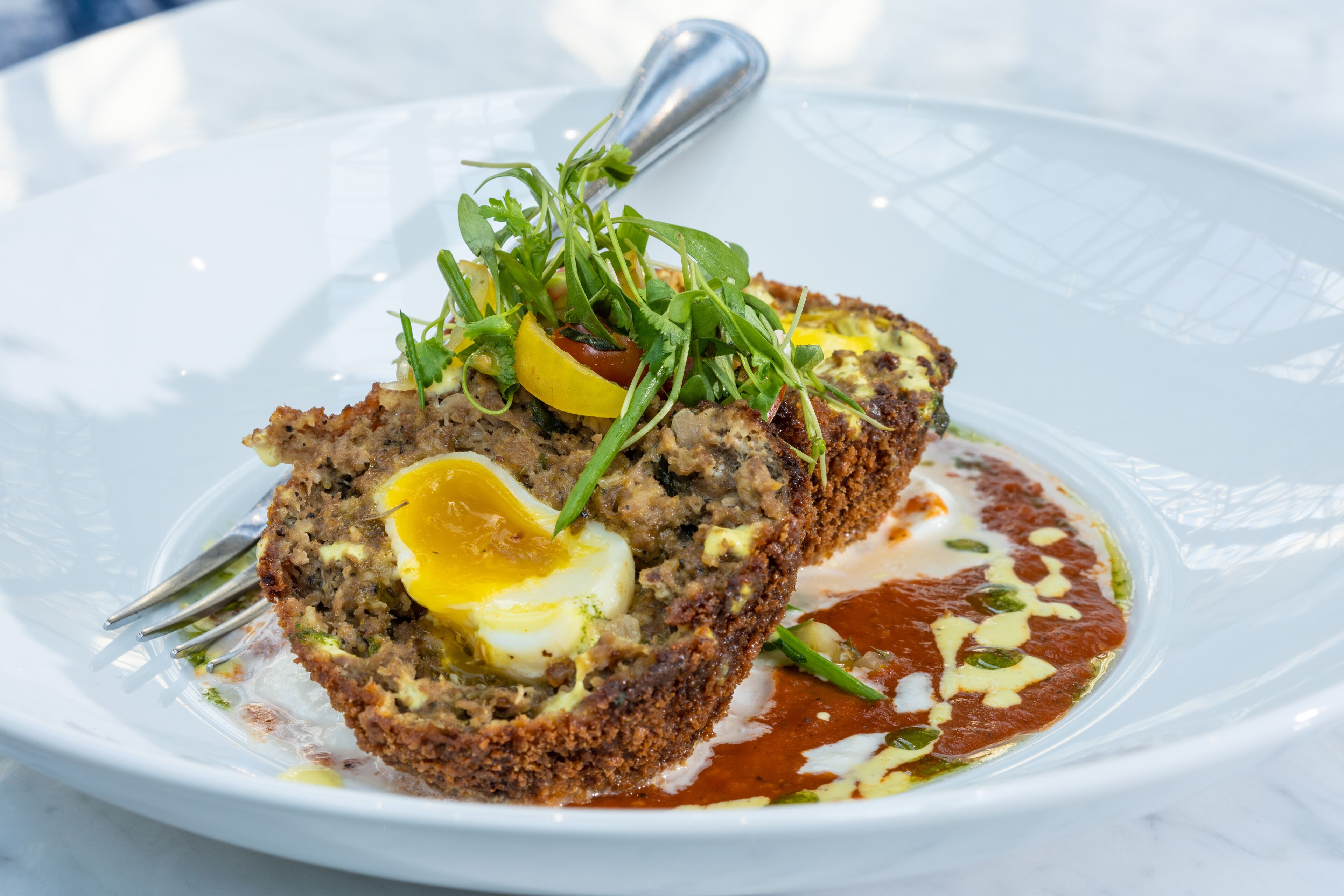 A dish with a slice of meat, egg, and greens on a white plate.
