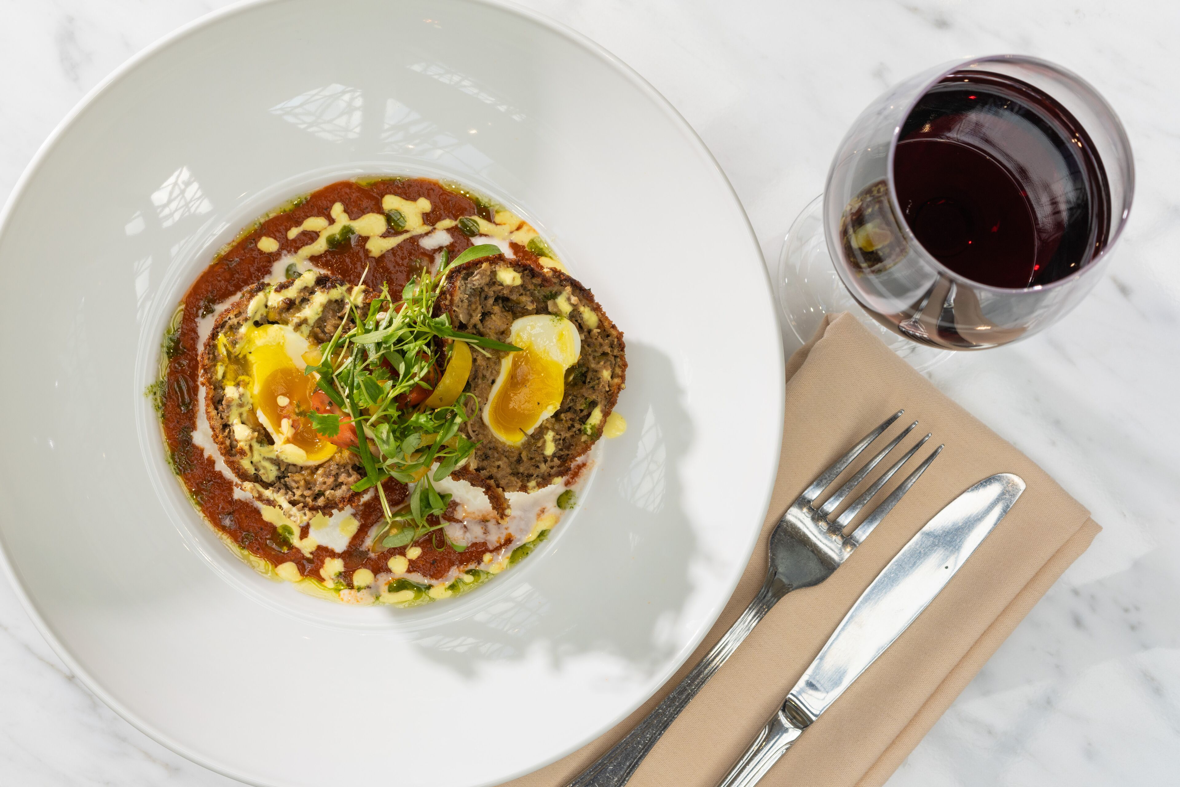 A white plate with a dish, a glass of red wine, and a fork and knife on a beige napkin.