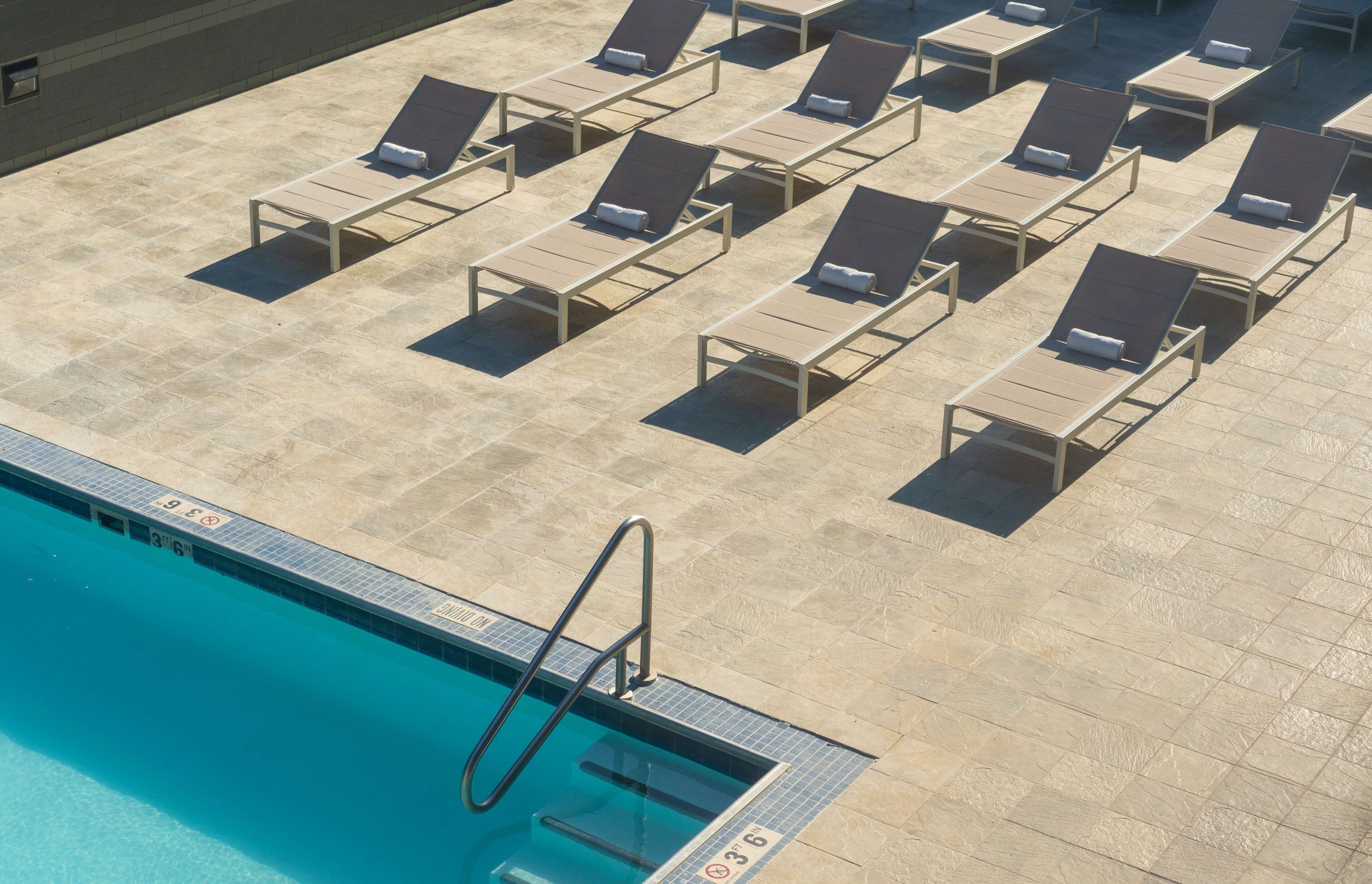 A poolside area with sun loungers arranged in rows, a pool with clear blue water, and a tiled deck.
