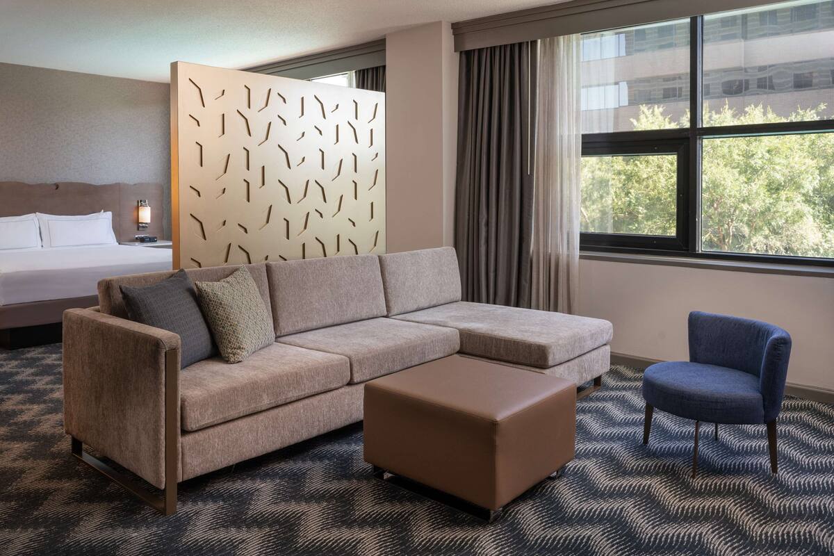 A hotel room with a large bed, a sofa, a chair, a patterned carpet, and a window with a view of trees.