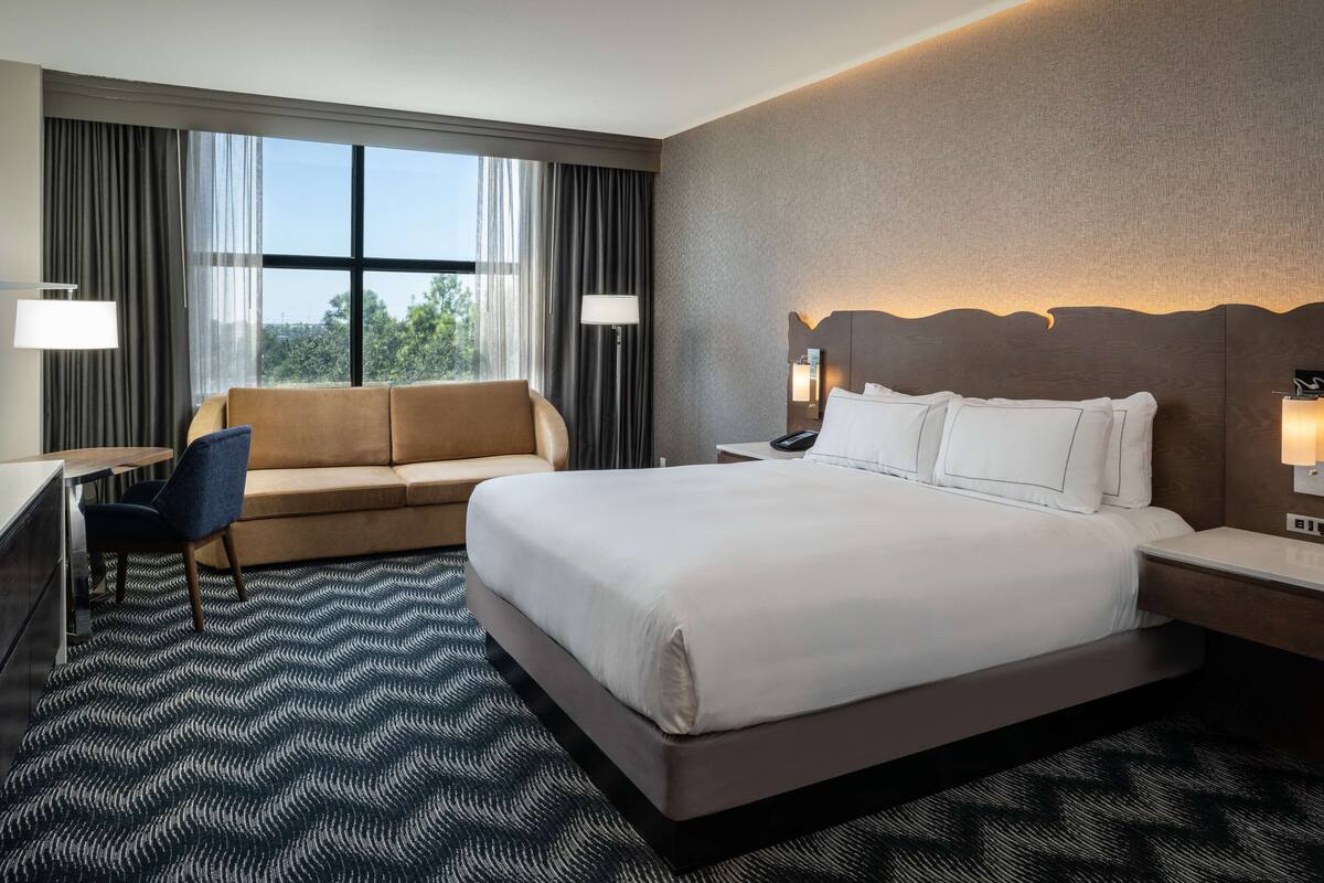 A hotel room with a large bed, a sofa, a desk, and a view of the outdoors through the window.