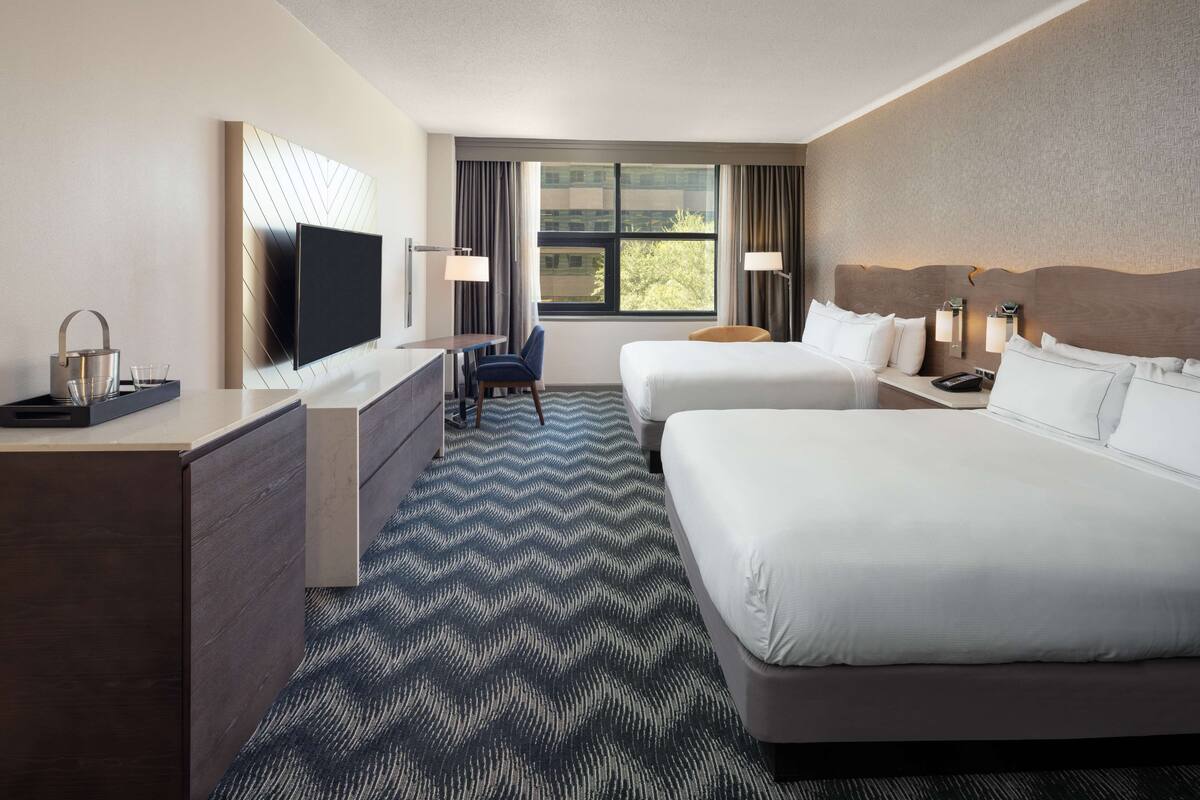A hotel room with two beds, a desk, a TV, and a window with curtains.