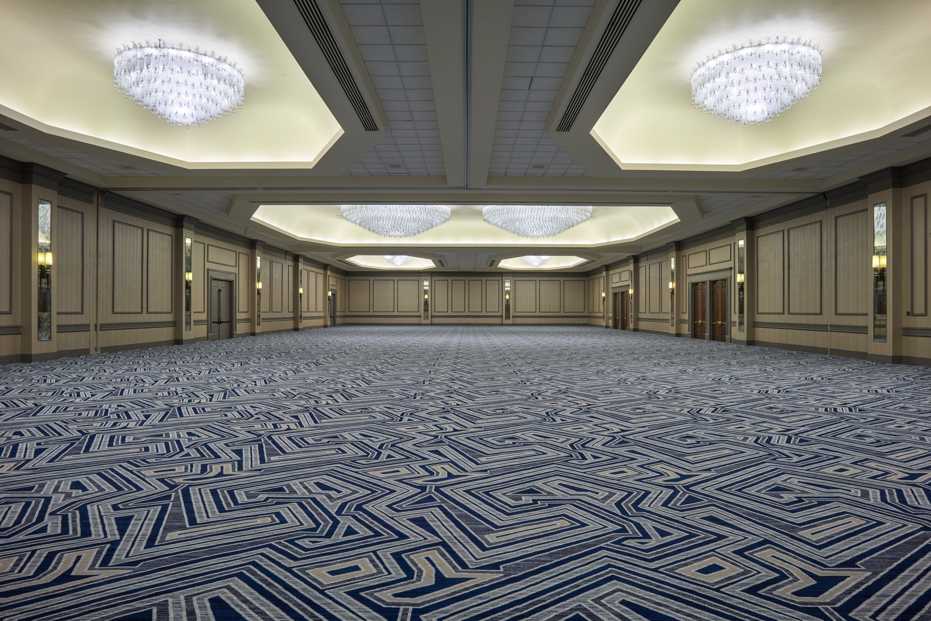 A spacious conference room with a geometric patterned carpet, recessed lighting, and a high ceiling with chandeliers.