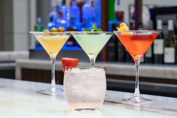 Tres vasos de martini sobre una barra con hielo, cada uno con una bebida de color diferente y adornos.