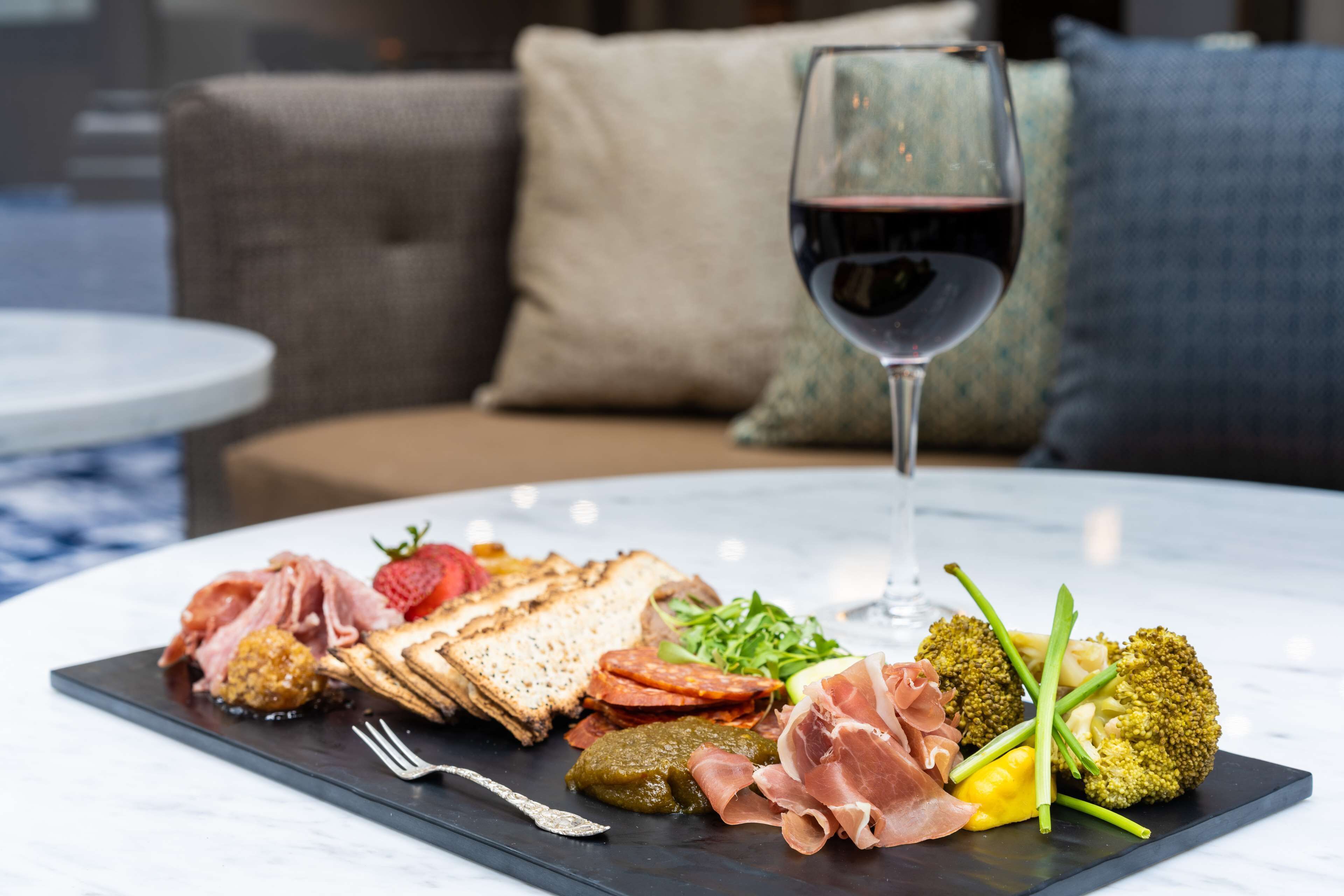 A charcuterie board with various meats, cheeses, and vegetables on a marble table, accompanied by a glass of red wine.