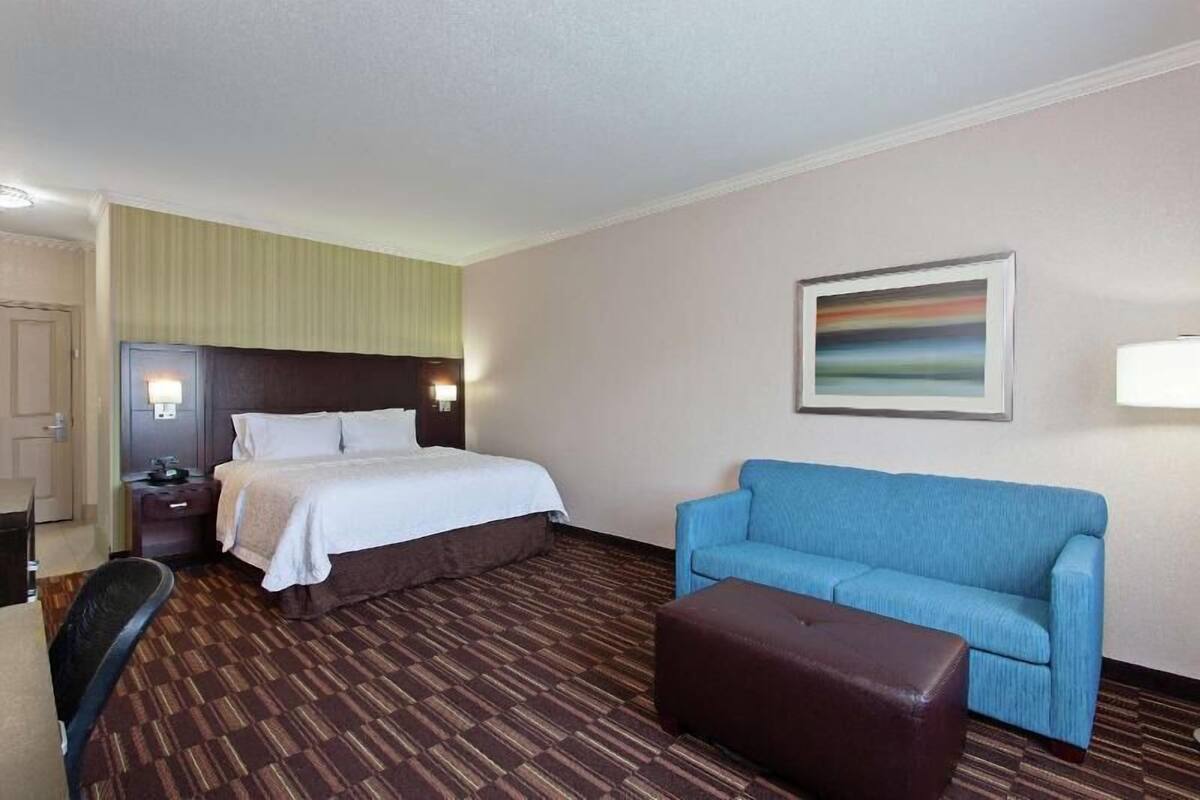 A hotel room with a bed, a blue sofa, a brown ottoman, a nightstand, and a painting on the wall.