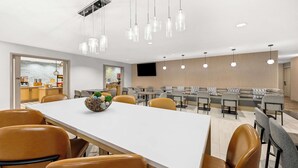 A modern dining area with a long table, multiple chairs, hanging pendant lights, and a television on the wall.
