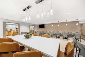A modern dining area with a long table, multiple chairs, hanging pendant lights, and a television on the wall.