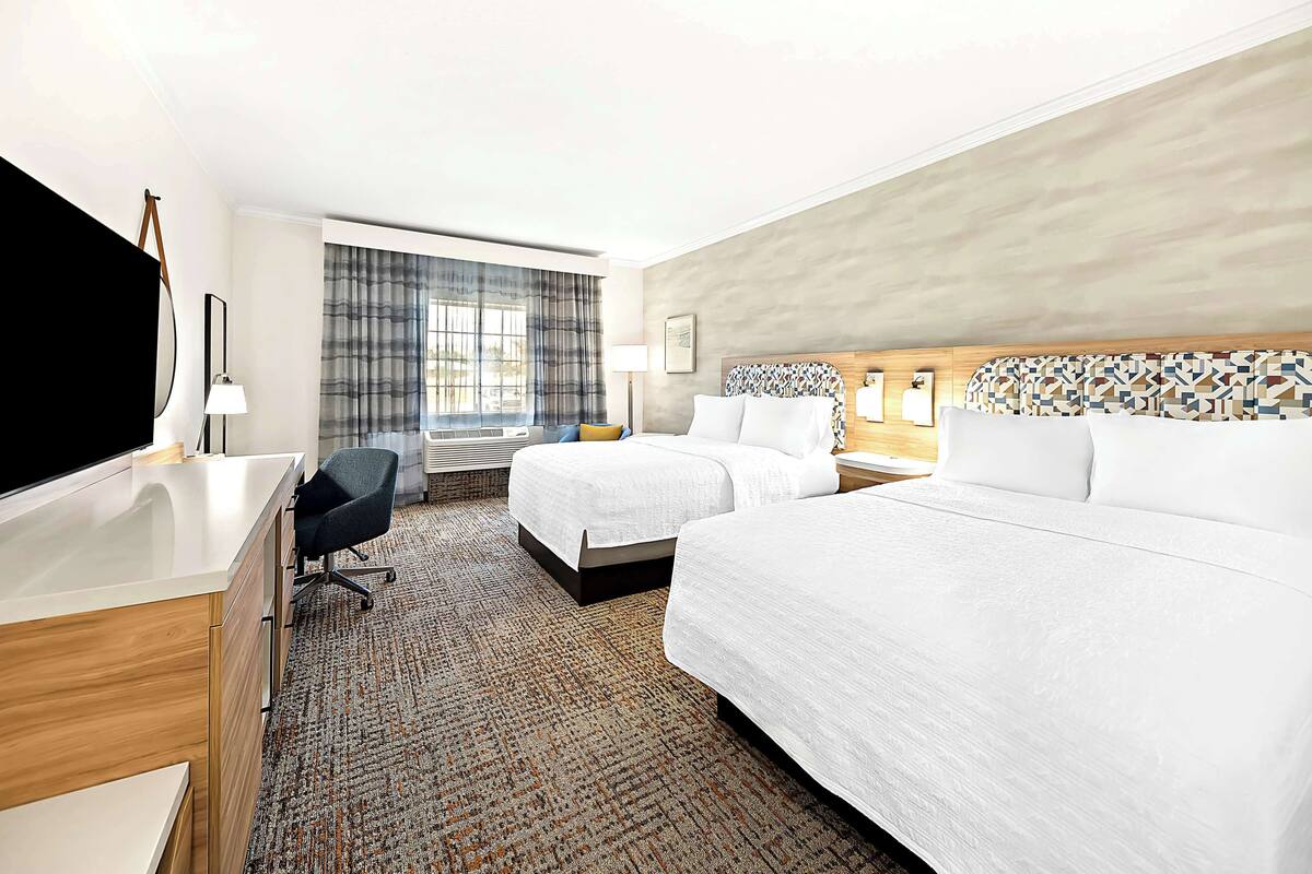 A hotel room with two beds, a desk, a chair, a television, and a large window with curtains.