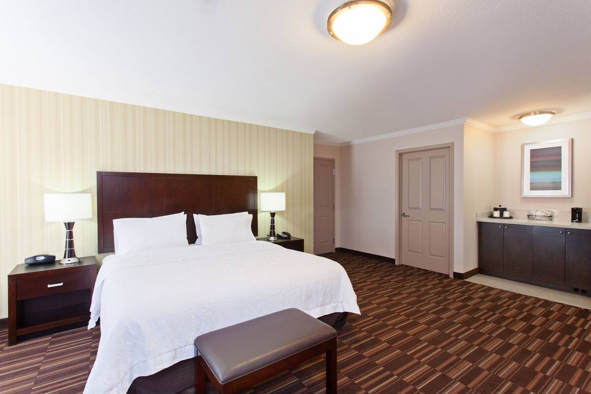 A hotel room with a large bed, bedside tables with lamps, a bench, a wardrobe, and a door.