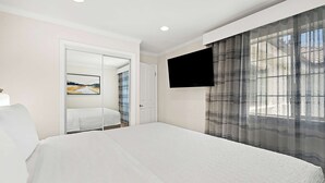 A hotel room with a bed, a TV mounted on the wall, a door leading to another room, and a window with sheer curtains.