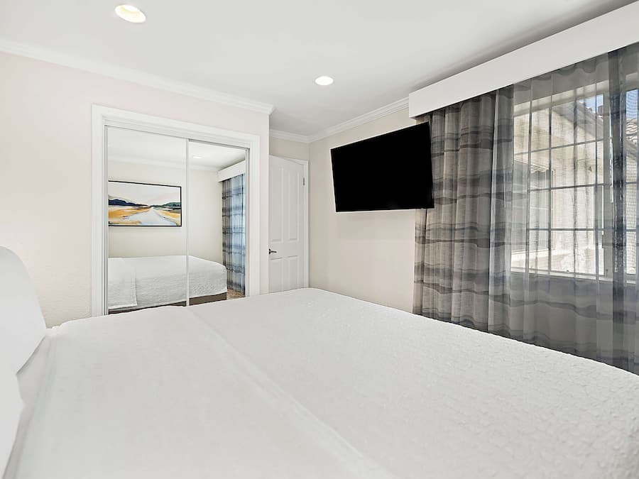 A hotel room with a bed, a TV mounted on the wall, a door leading to another room, and a window with sheer curtains.
