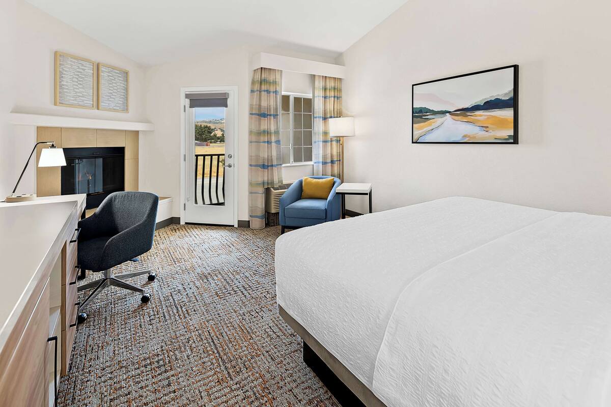 A hotel room with a bed, a desk, a chair, a fireplace, a window with curtains, and a painting on the wall.