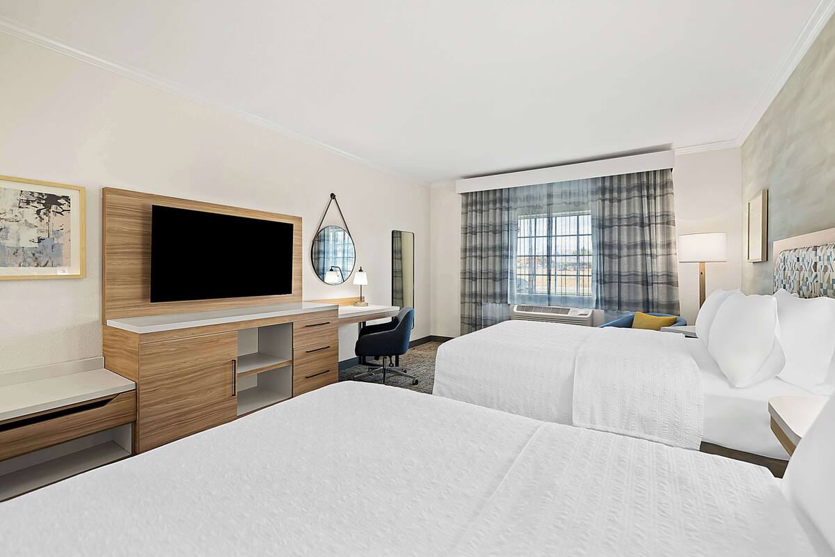 A hotel room with two beds, a desk with a flat-screen TV, a chair, a lamp, and a large window with curtains.