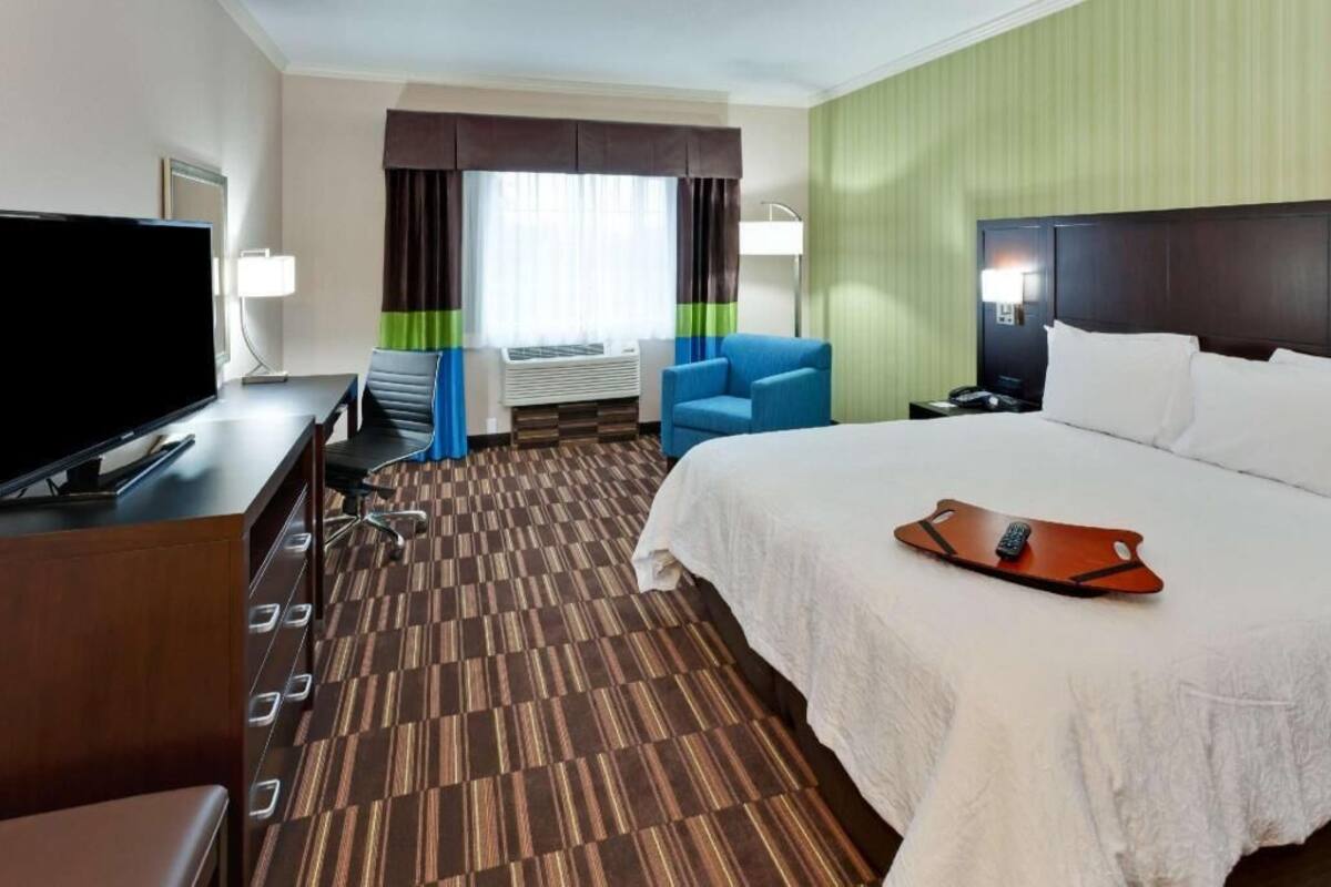 A hotel room with a bed, a desk with a TV, a chair, a desk lamp, a bedside table, and a window with curtains.