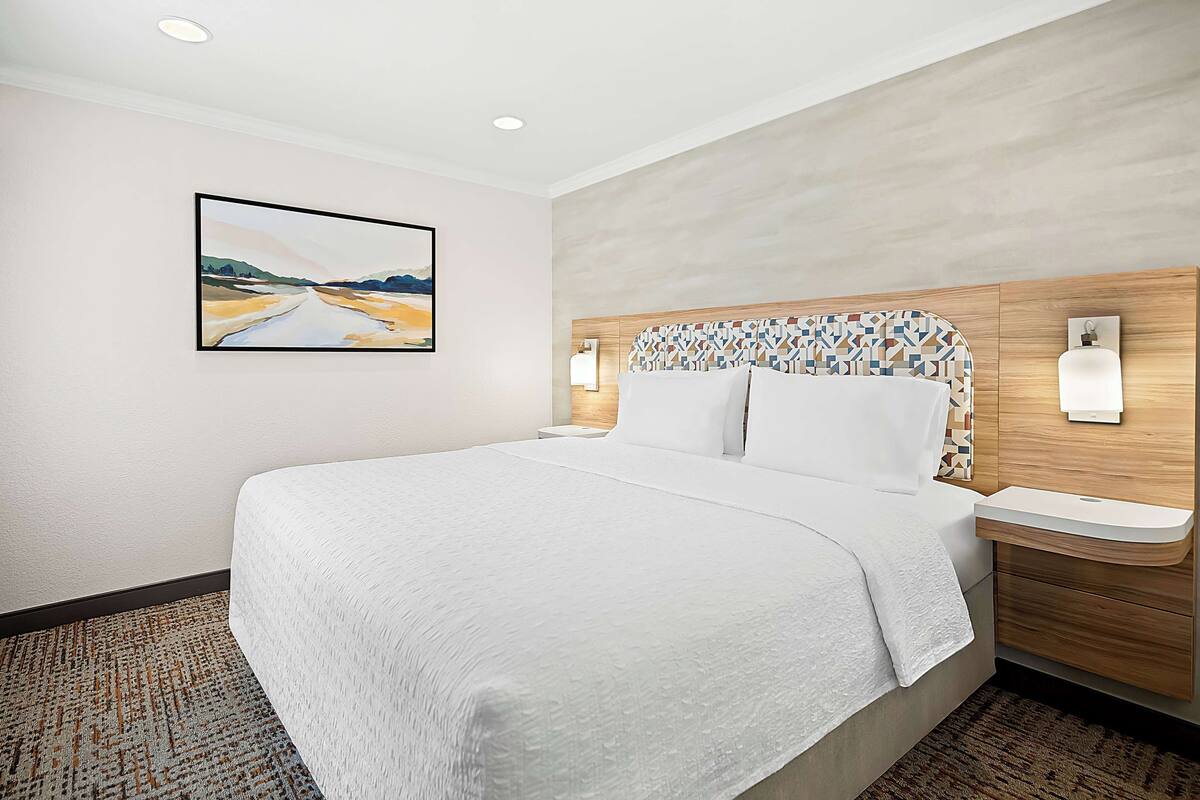 A hotel room with a large bed, a headboard with a geometric pattern, a painting on the wall, and a nightstand with a lamp.
