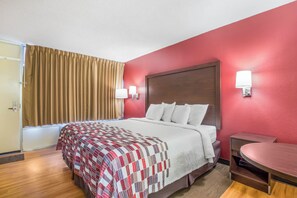 Habitación de hotel con cama, mesita de noche, una silla, ventana con cortinas y una lámpara de pared.
