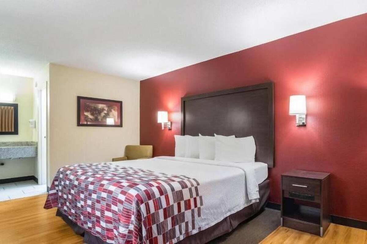 A hotel room with a bed, a nightstand, a lamp, a wall-mounted light, and a framed picture on the wall.