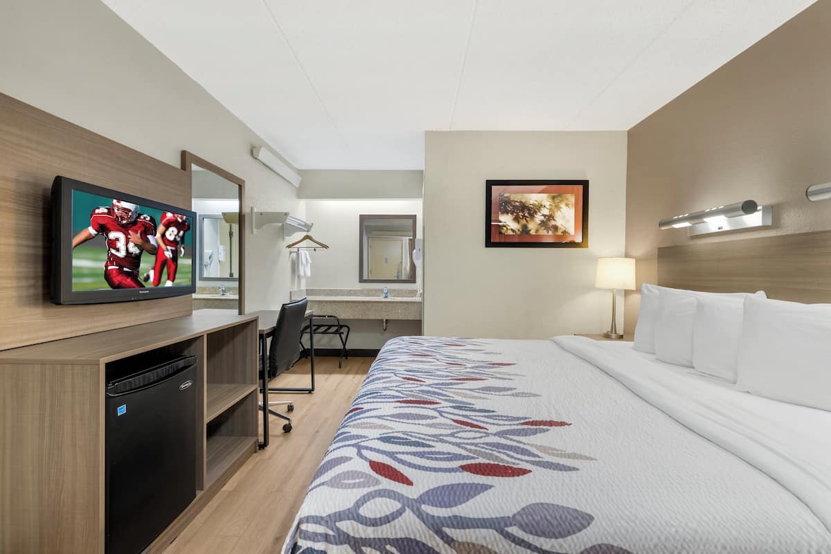 Ein Hotelzimmer mit einem Bett, einem Fernseher auf einer Holzkommode, einem Schreibtisch mit Stuhl, einer Schreibtischlampe und einem Bild an der Wand.