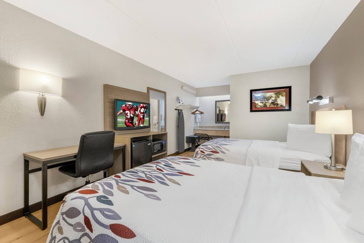 Une chambre d’hôtel avec un bureau, deux lits, une télévision, un tableau et une lampe.
