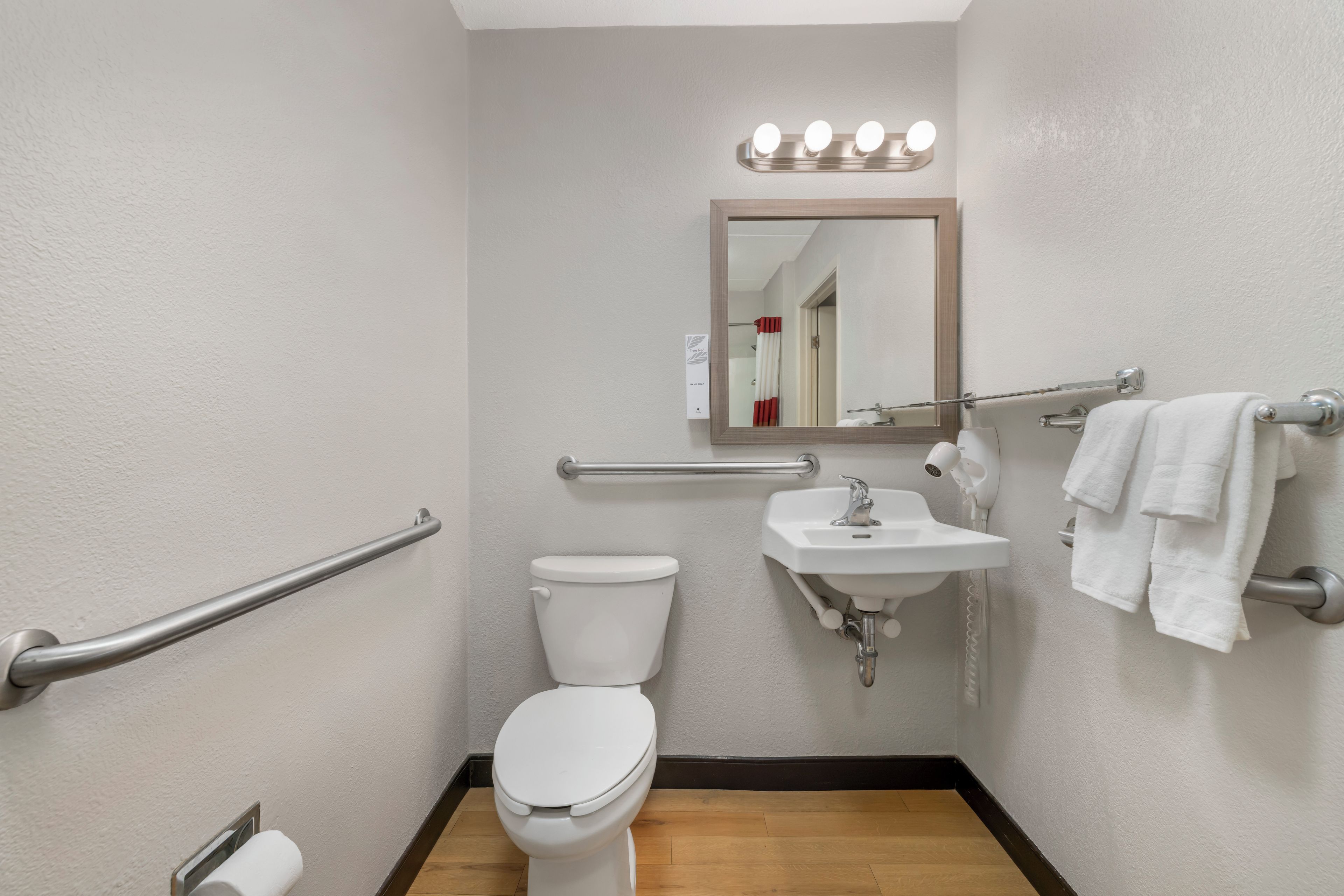 A standard hotel bathroom with a toilet, sink, handrails, a mirror with lights, and a towel rack.