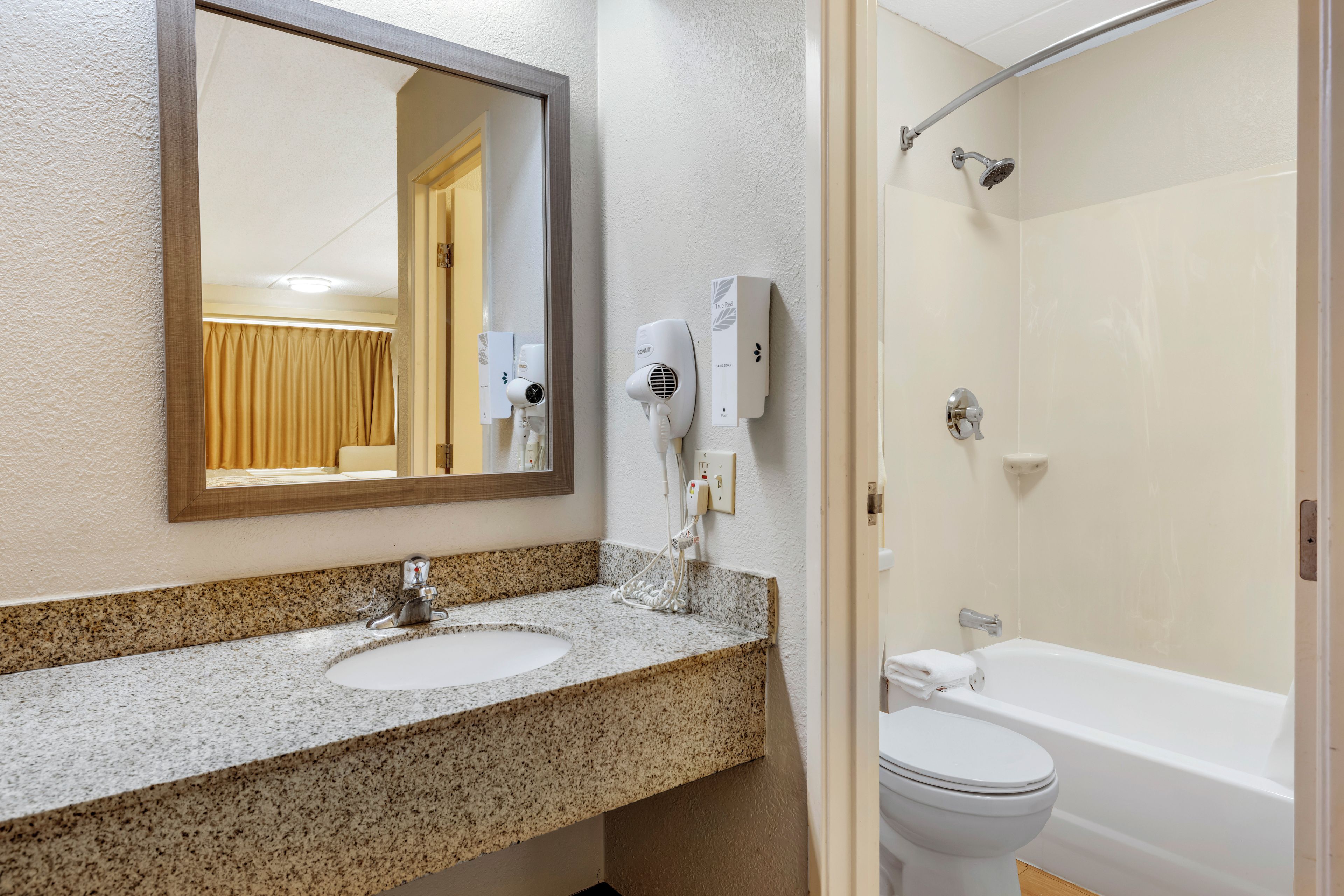 A bathroom with a granite countertop sink, a mirror, a handheld hair dryer, a handheld soap dispenser, and a bathtub with a showerhead.