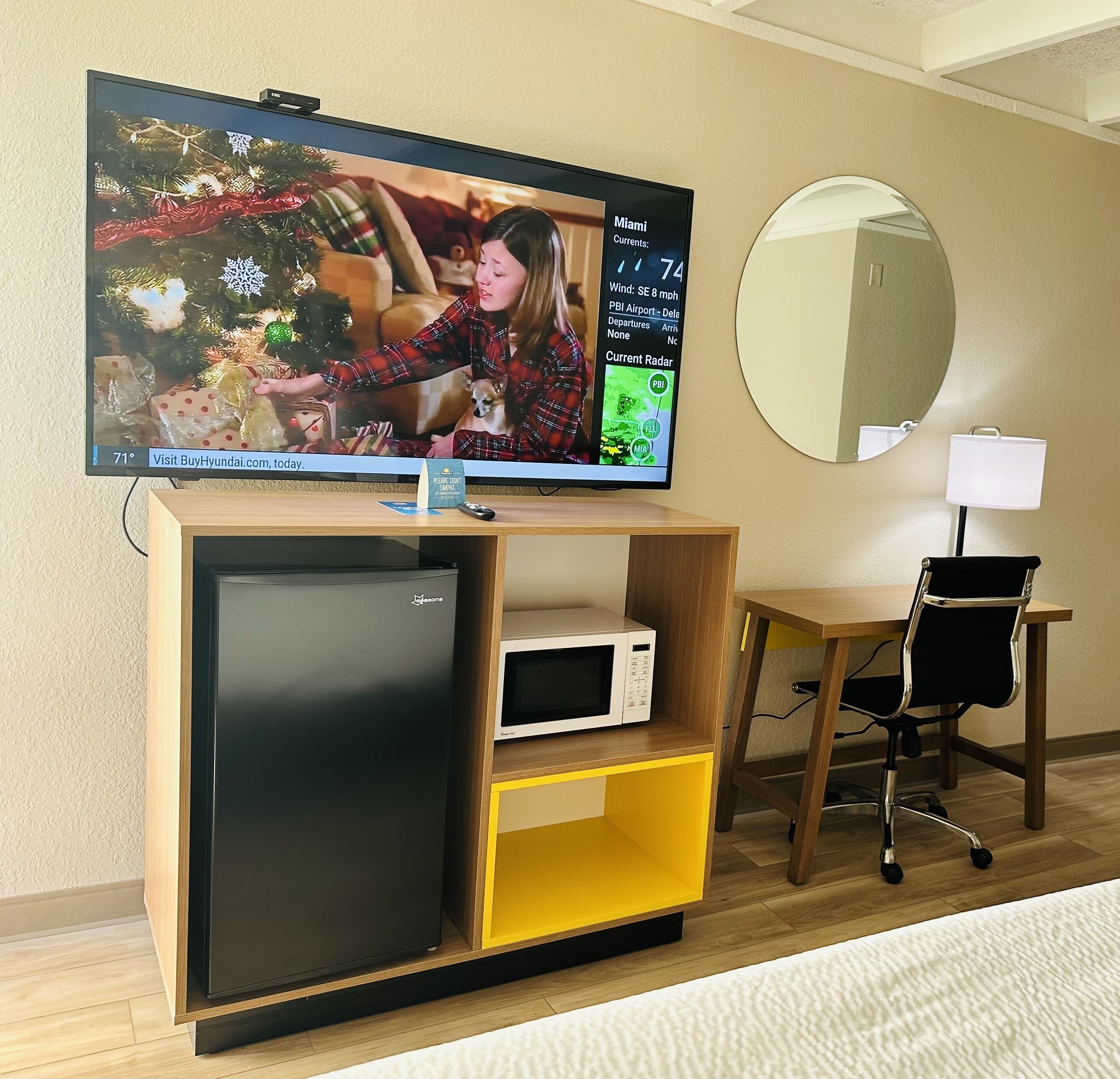 Habitación de hotel con televisión de pantalla plana montada en un mueble de madera, microondas, refrigerador pequeño, escritorio con silla y espejo redondo en la pared.
