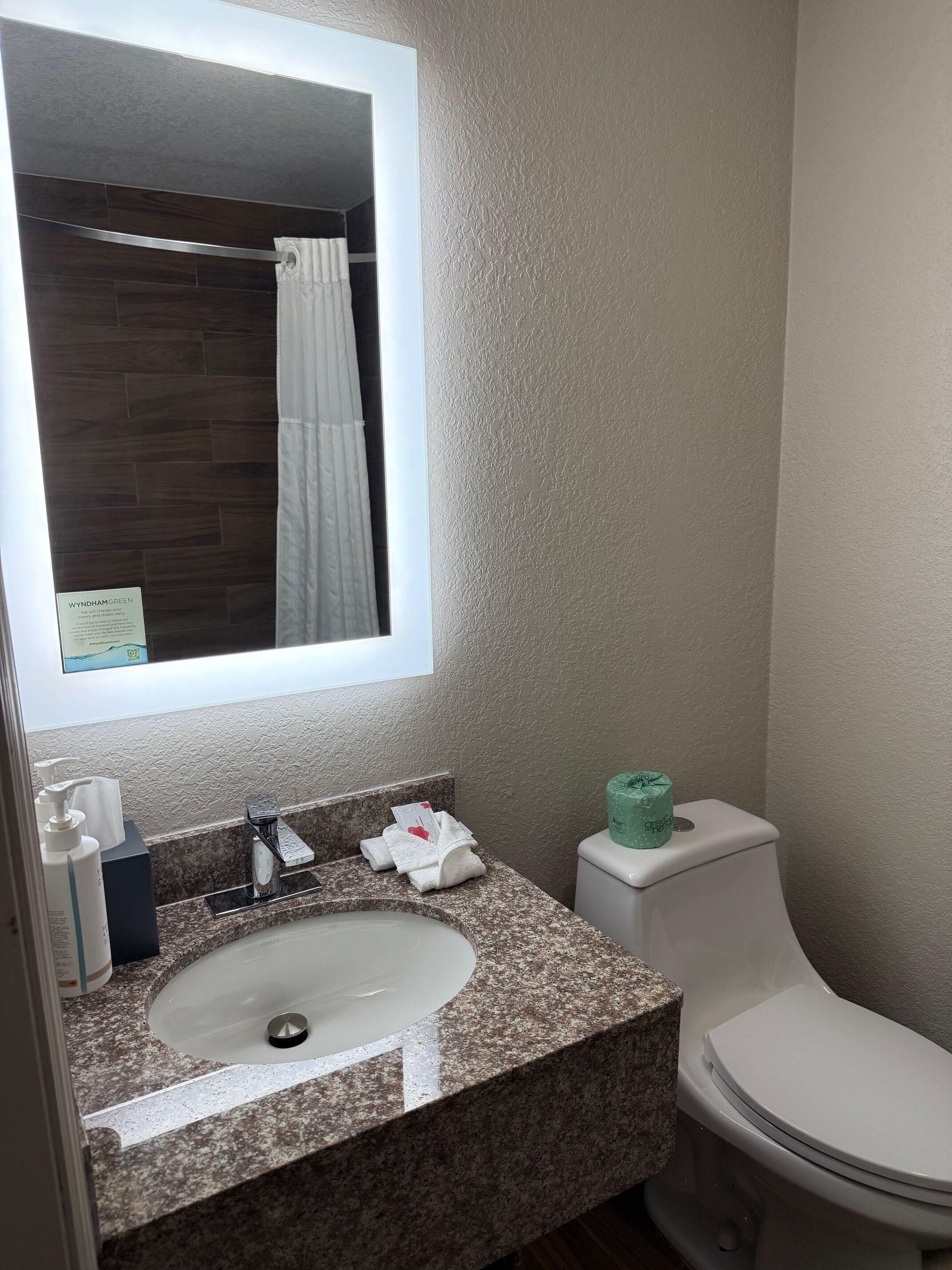 Une salle de bain avec un comptoir en granit, un lavabo blanc, un miroir, un distributeur de serviettes et des toilettes.