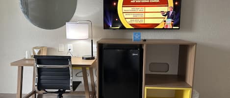 Una habitación con escritorio, silla, computadora, televisor y un refrigerador pequeño.