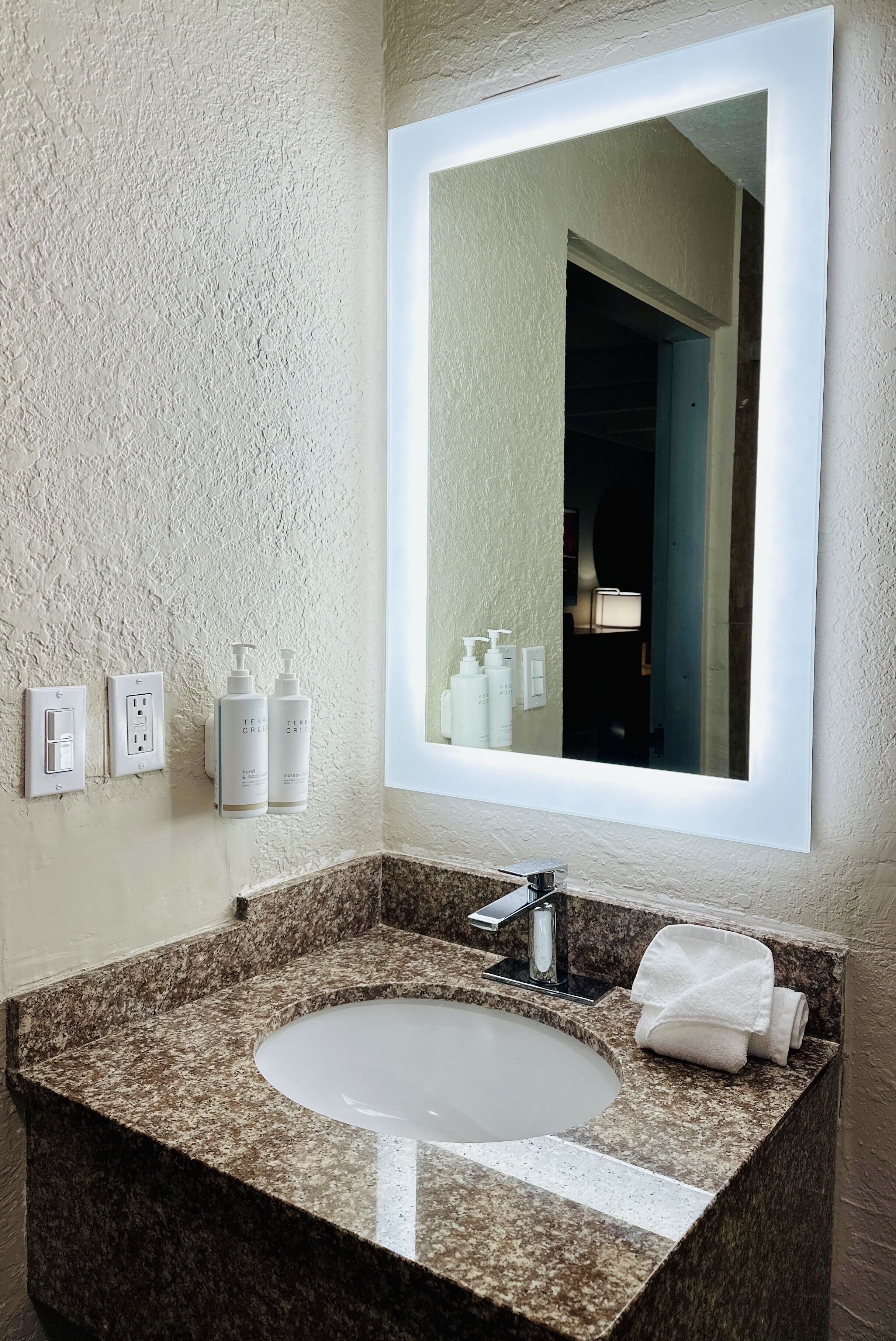 A bathroom with a granite countertop sink, a large mirror, and two bottles of toiletries on the wall.