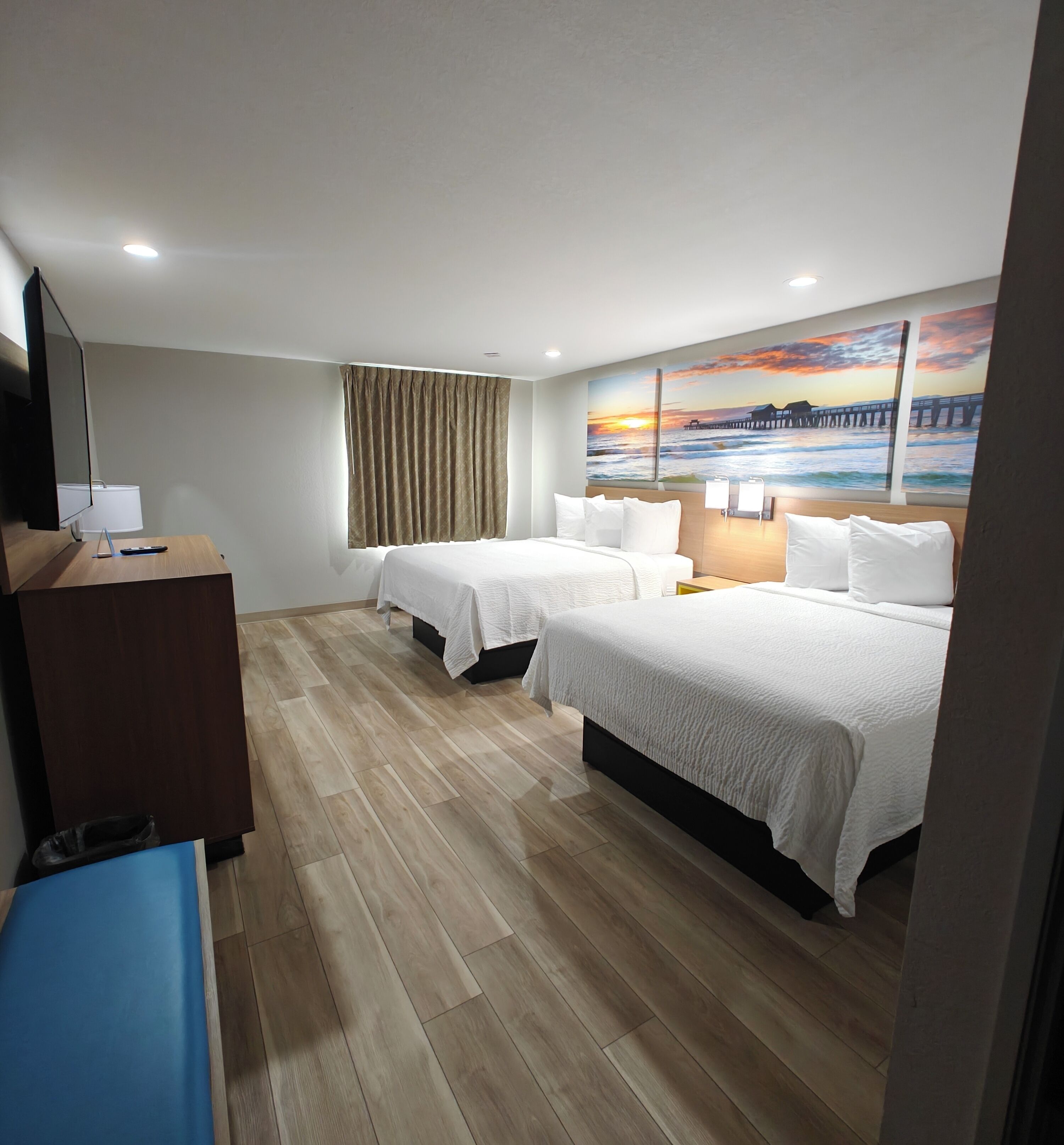 A hotel room with two beds, a desk, a TV, and a painting of a sunset over the ocean.