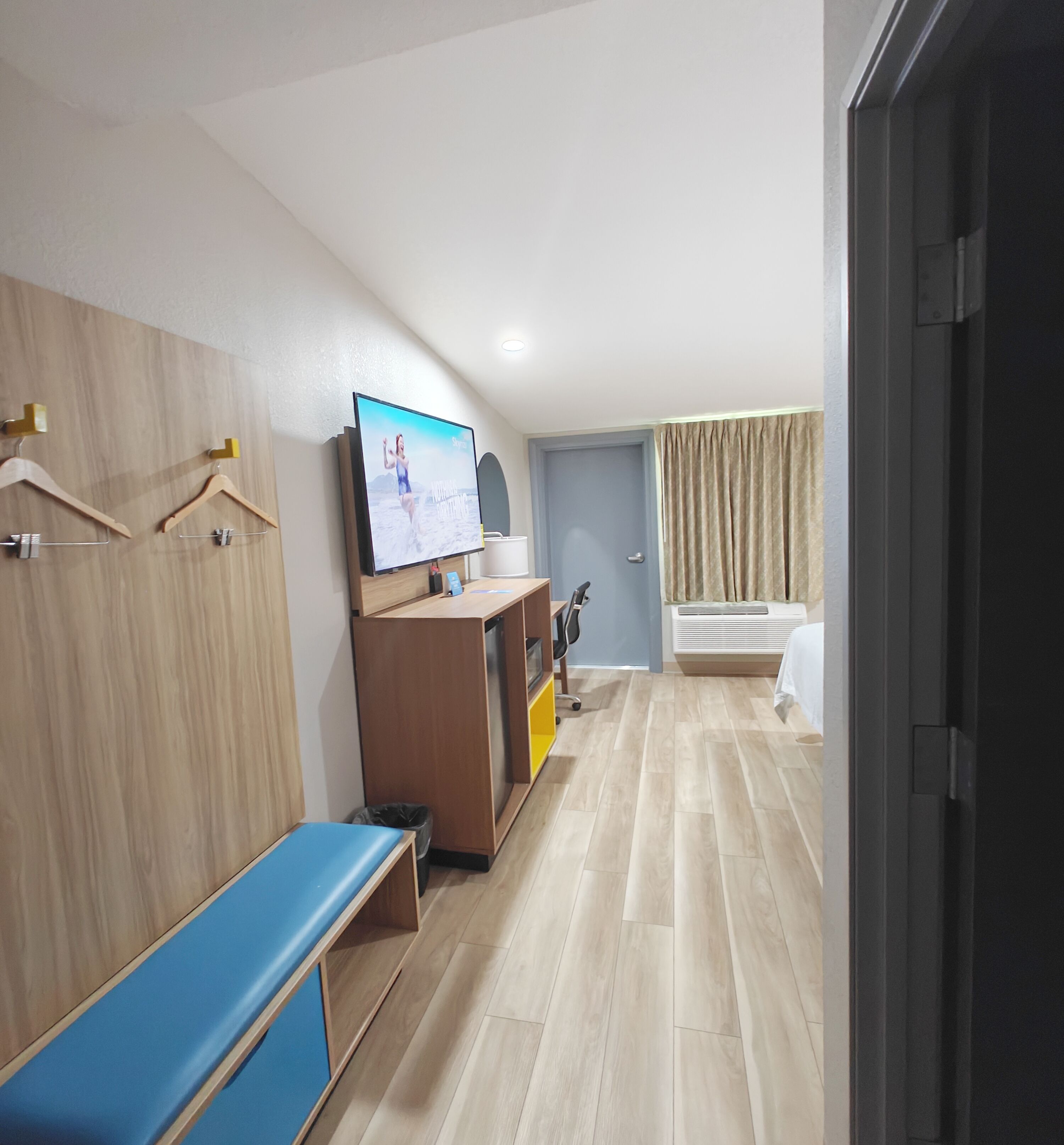 A modern hotel room with a wooden wardrobe, a television, a desk with a computer, a bench, and a bathroom with a curtain.