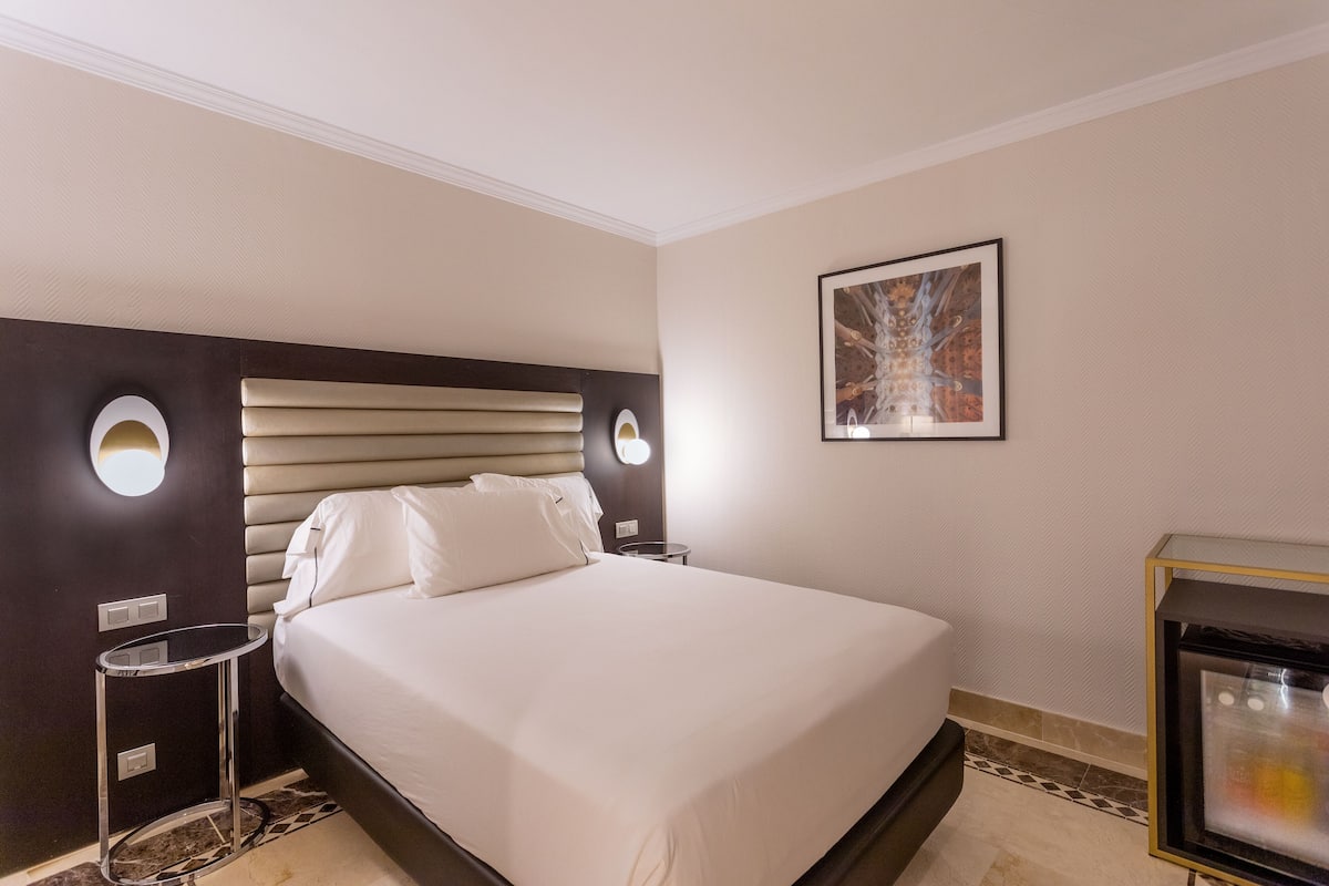 A hotel room with a large bed, two bedside tables, a wall-mounted artwork, and a small TV on a stand.