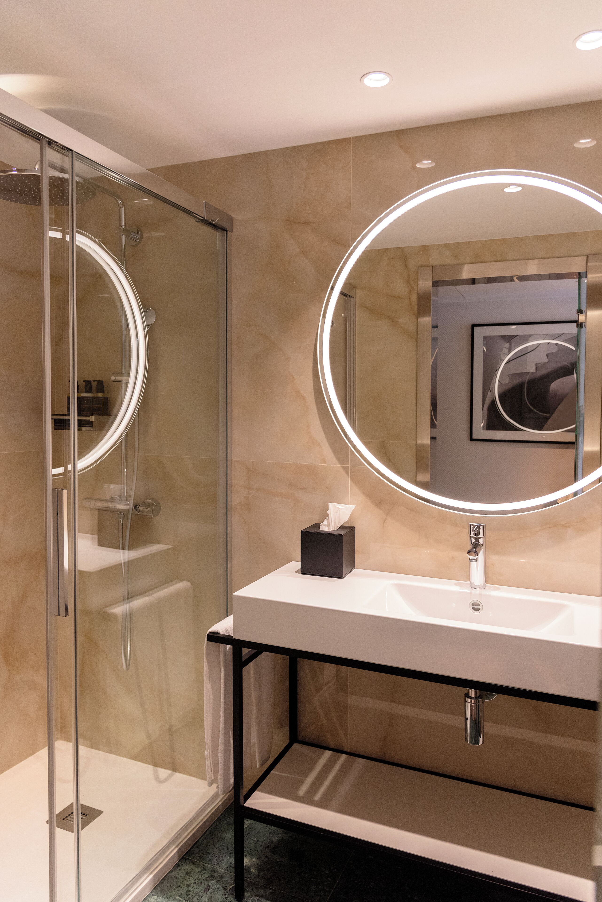 Un baño moderno con ducha de vidrio, un espejo redondo y un lavamanos blanco con marco negro.