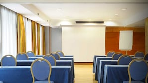 Un salón de conferencias con filas de mesas y sillas, una pizarra y una pantalla de proyección.