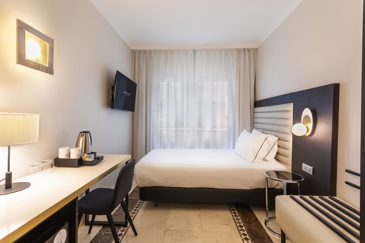 A hotel room with a bed, a desk with a lamp, a chair, a TV mounted on the wall, and a window with sheer curtains.