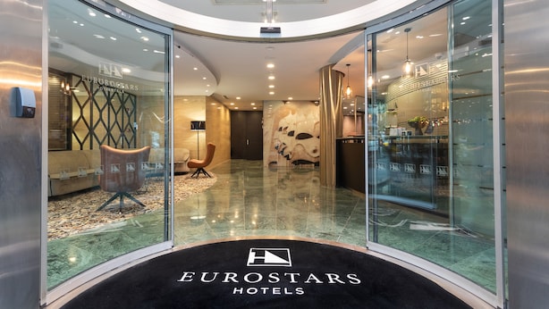 Un vestíbulo de hotel moderno con paredes de vidrio, un área de estar central y una recepción visible.
