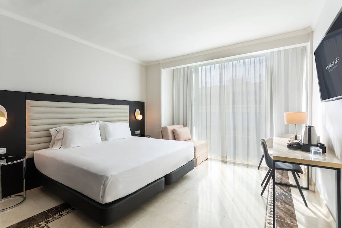 A hotel room with a large bed, a desk with a chair, a TV, and a window with curtains.