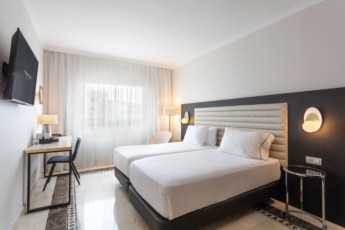 A hotel room with two beds, a desk, a chair, a TV, and a lamp.