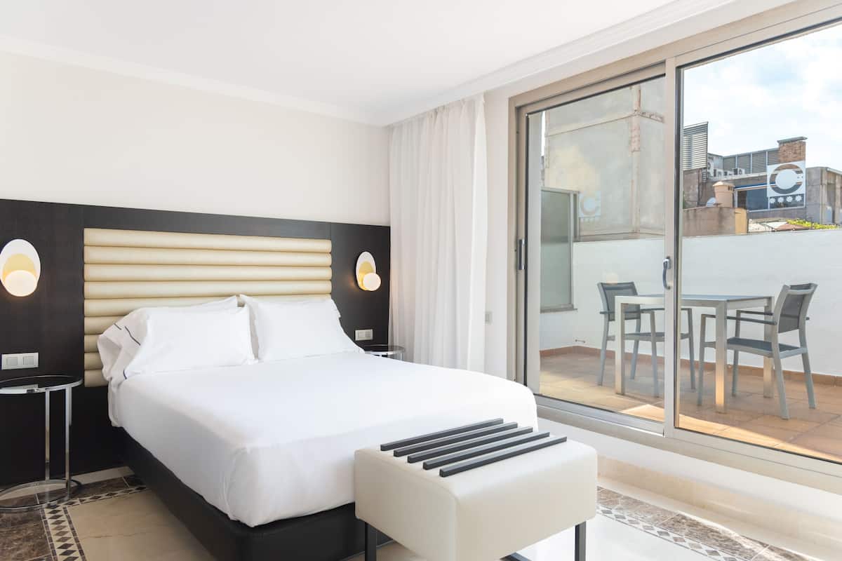 A hotel room with a large bed, a small bedside table, a chair, and a view of the outdoor patio.