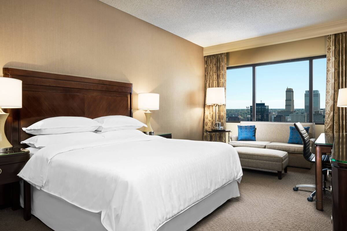 A hotel room with a large bed, two bedside tables with lamps, a sofa, and a view of the city skyline.