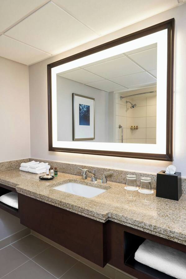 A modern bathroom with a large mirror, granite countertop, dual sinks, and a shower visible in the reflection.