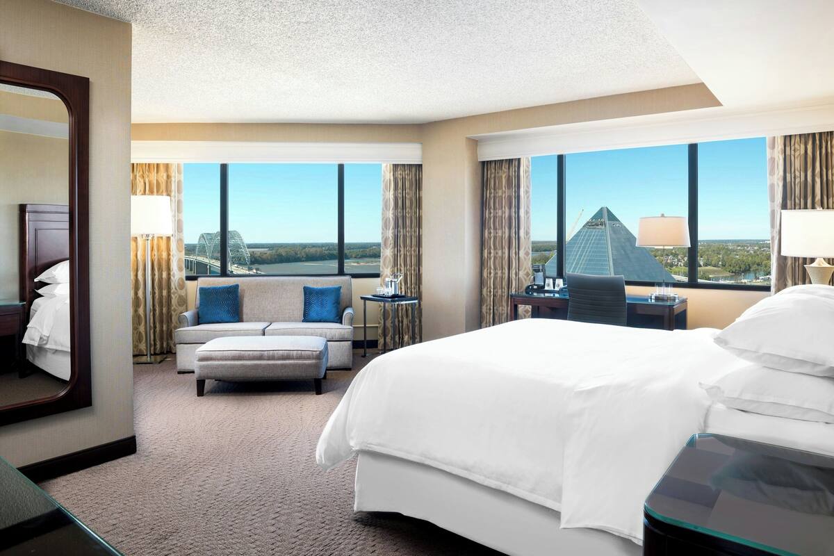 A hotel room with a large bed, a sofa, a desk, and a view of a bridge and pyramid.